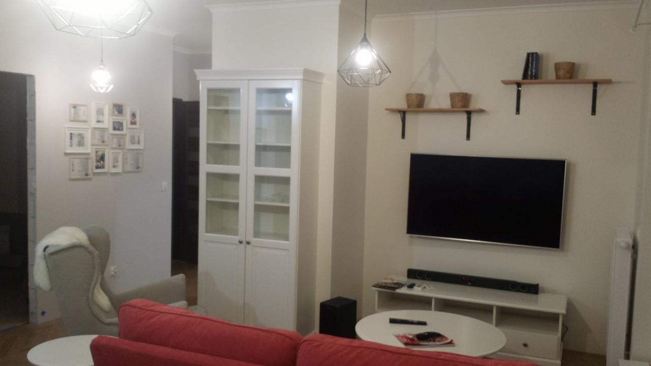 Jasne wnętrze salonu z białą szafą przeszkloną, telewizorem na ścianie, drewnianymi półkami z dekoracjami i czerwoną sofą na pierwszym planie.