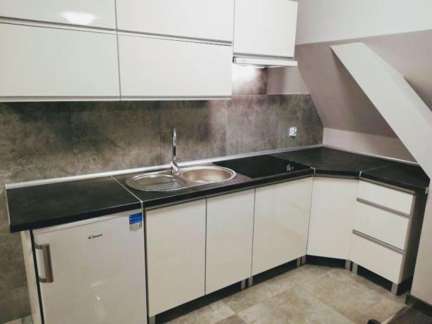 Jasna kuchnia z białymi szafkami i czarnym blatem, zlew dwukomorowy i płyta grzewcza, ukośny sufit w tle.