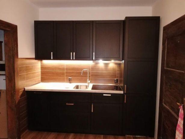 Ciemna zabudowa kuchenna z górnymi szafkami, blatem z umywalką i płytą grzewczą oraz podświetleniem LED podszafkowym na tle płytek imitujących drewno.