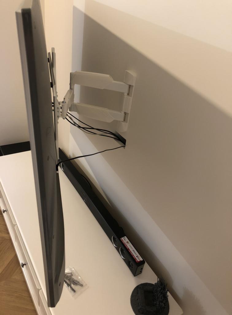 Telewizor zamontowany na ścianie za pomocą białego, regulowanego uchwytu, widoczne kable, listwa głośnikowa i akcesoria montażowe na białej komodzie.