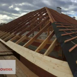 Konstrukcja drewniana dachu w trakcie budowy, widoczne elementy więźby, membrana dachowa częściowo rozłożona, rusztowanie wzdłuż krawędzi, budynek w stanie surowym otwartym, logo firmy w lewym...