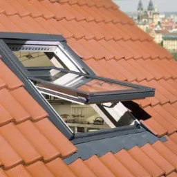 Otwarty okno dachowe z szarą ramą na dachu pokrytym czerwoną dachówką; w tle zabudowania miejskie z wieżami.