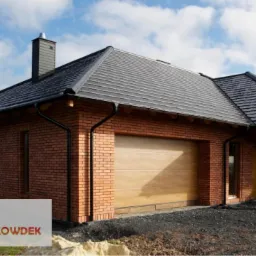 Dom jednorodzinny z cegły klinkierowej z ciemnoszarą dachówką ceramiczną, rynnami w kolorze czarnym i drewnianą bramą garażową, na tle błękitnego nieba z chmurami. Widoczny komin z tego samego...