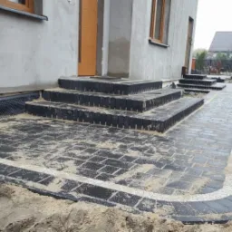 Ułożona kostka brukowa w kolorze grafitowym z białym obramowaniem, prowadząca do schodów wejściowych domu. Widoczna skrzynka gazowa na elewacji oraz rozsypany piasek na powierzchni.