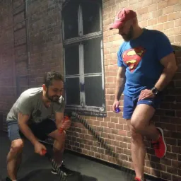 Mężczyzna ćwiczy z liną treningową pod okiem trenera w koszulce Supermana na tle ściany z cegieł z namalowanym oknem.