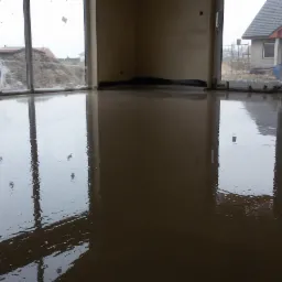 Świeżo wylana, gładka wylewka anhydrytowa w pomieszczeniu z dużymi oknami i widokiem na plac budowy, widoczna rura wentylacyjna z niebieską nasadką.