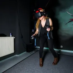 Kobieta w Łodzi używa zestawu VR, trzymając kontrolery w ciemnym pomieszczeniu z dekoracją na ścianie.