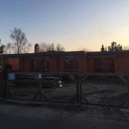 Alex Sp zoo - Budowa domu jednorodzinnego z czerwonej cegły, widok zza drewnianego ogrodzenia wczesnym rankiem, widoczne otwory okienne i kominy.