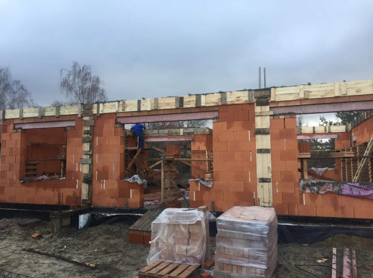 Wznoszenie ścian domu z czerwonej cegły ceramicznej Porotherm, widoczne rusztowanie i pracownik w niebieskim ubraniu, drewniane szalunki nad otworami okiennymi, palety cegieł w folii na placu budowy.