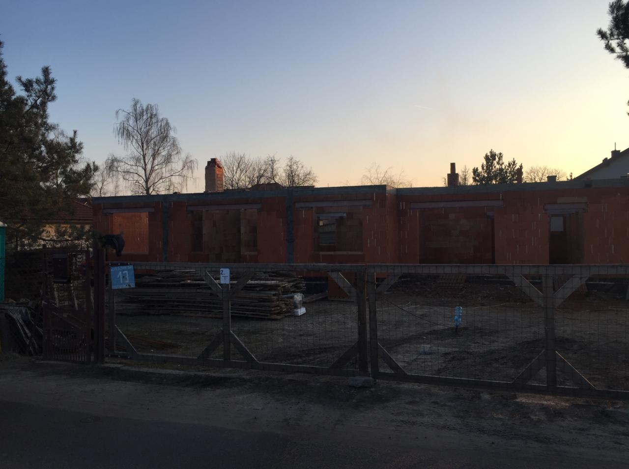 Budowa domu jednorodzinnego z czerwonej cegły, widok zza drewnianego ogrodzenia wczesnym rankiem, widoczne otwory okienne i kominy.