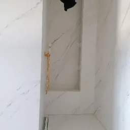 Wykończona łazienka z białymi marmurowymi płytkami, złotą armaturą prysznicową i wnęką na kosmetyki.