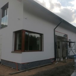 Przeszon - Nowoczesny dom jednorodzinny w trakcie prac elewacyjnych: ocieplenie styropianem, cokół z szarej cegły klinkierowej, widoczne rusztowanie i paleta z kostką brukową. Pracownik w żółtej koszulce...
