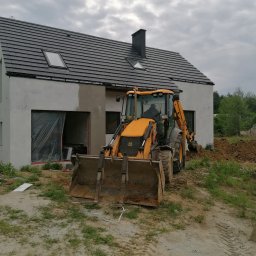 Przeszon - Żółta koparko-ładowarka JCB na tle surowego domu jednorodzinnego z szarą dachówką i częściowo otynkowaną elewacją, okna zabezpieczone folią.