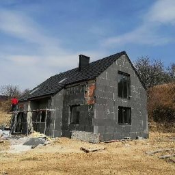 Przeszon - Dom jednorodzinny w trakcie ocieplania elewacji zewnętrznej styropianem, widoczne rusztowania, pracownik w czerwonej koszuli na rusztowaniu, częściowo odsłonięta cegła.