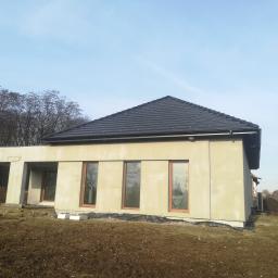 Przeszon - Nowoczesny dom jednorodzinny w trakcie budowy, otynkowany na szaro, z ciemną dachówką, widoczne okna z brązowymi ramami i przygotowany teren wokół budynku.