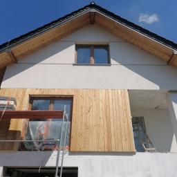 Przeszon - Dom jednorodzinny w trakcie montażu drewnianej elewacji, widoczne rusztowanie z materiałami budowlanymi oraz niezabezpieczone okno folią.