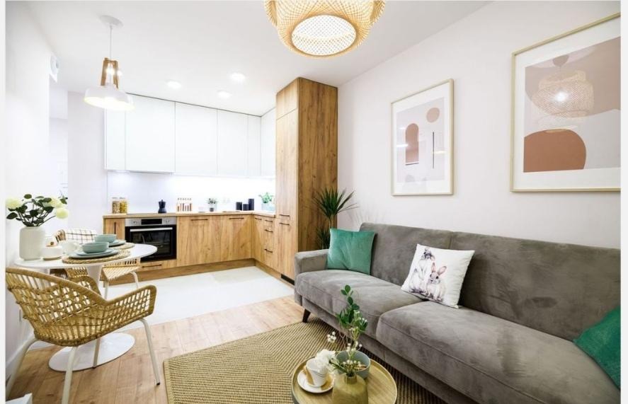 Jasne mieszkanie z aneksem kuchennym: drewniane szafki, białe fronty, okrągły stół z plecionymi krzesłami, szara sofa z poduszkami i abstrakcyjne obrazy na ścianie.