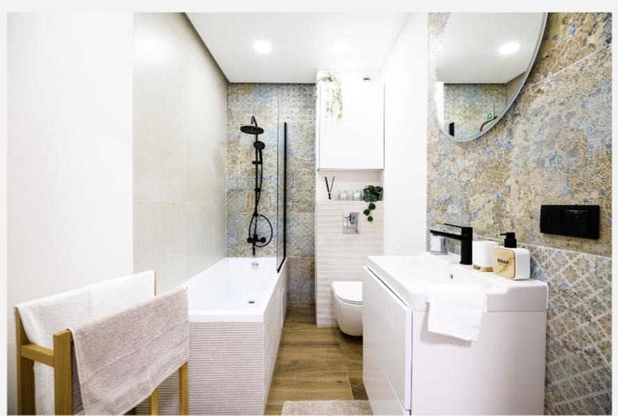 Nowoczesna łazienka z wanną i czarną armaturą, beżowo-niebieskie płytki na ścianach, drewniany regał na ręczniki, biała umywalka z dozownikiem mydła.