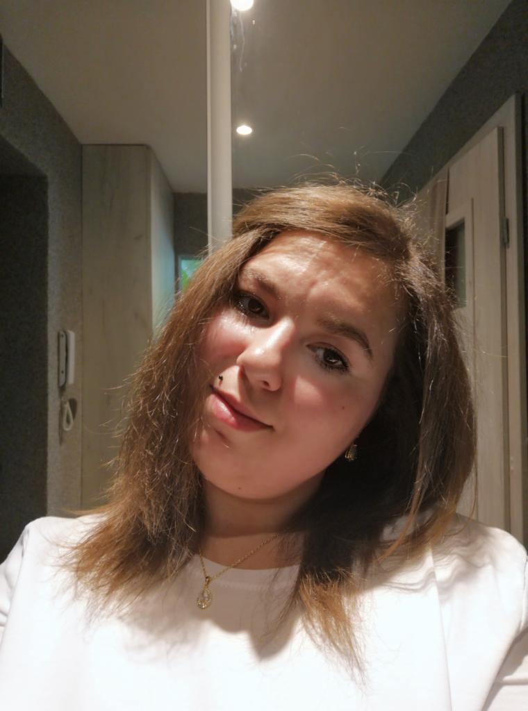 Portret kobiety z brązowymi włosami i złotym łańcuszkiem na tle wnętrza z oświetleniem punktowym. Uśmiech i delikatny makijaż.