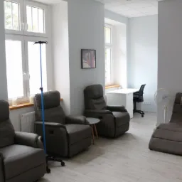 Wnętrze gabinetu z trzema rozkładanymi fotelami, stolikami, kroplówkami i biurkiem w tle, utrzymane w jasnej kolorystyce.