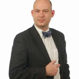 Mężczyzna w ciemnej marynarce i muszce, z ręką włożoną w połowę marynarki, pozuje na białym tle.