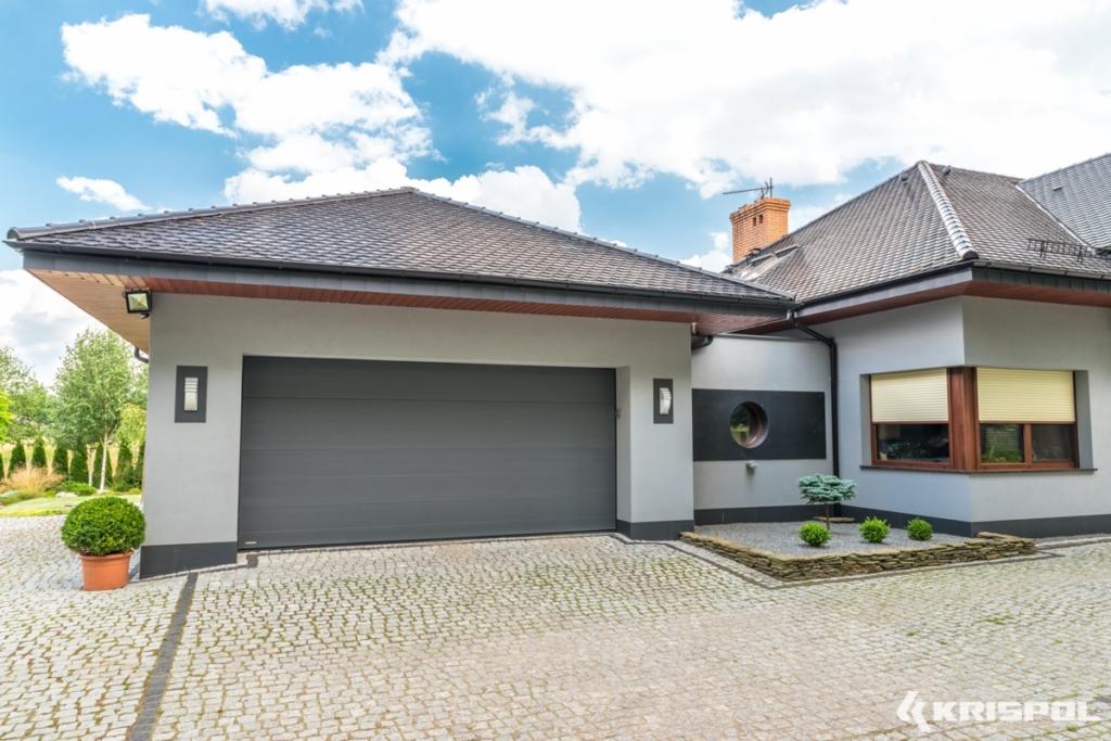Elegancki dom jednorodzinny z szarą bramą garażową segmentową i brukowanym podjazdem z kostki granitowej w słoneczny dzień.