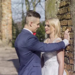 Para młoda w plenerze przy ceglanej ścianie; panna młoda w białej sukni, pan młody w granatowym garniturze z czerwoną muchą, oboje patrzą na siebie z uśmiechem.