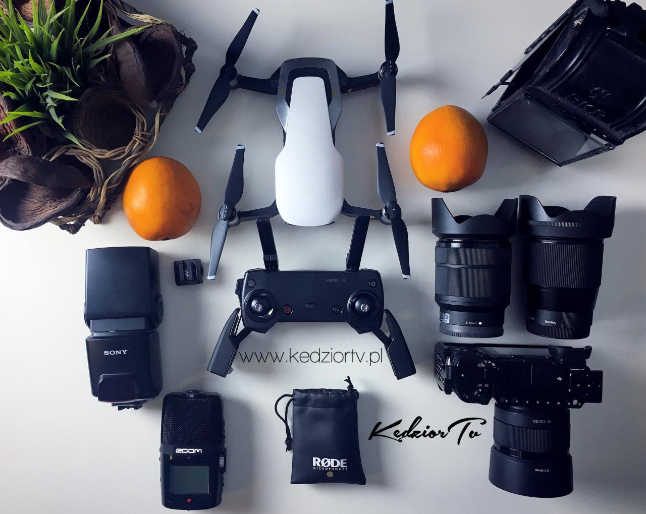 Dron Mavic Air, kontroler, kamera Sony, mikrofon Zoom, lampa błyskowa Sony, dwa obiektywy Sigma, torba Rode i dwie pomarańcze ułożone na białym tle, dekoracyjne gałązki w rogu.