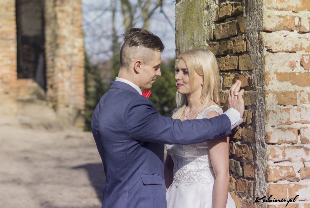Para młoda w plenerze przy ceglanej ścianie; panna młoda w białej sukni, pan młody w granatowym garniturze z czerwoną muchą, oboje patrzą na siebie z uśmiechem.