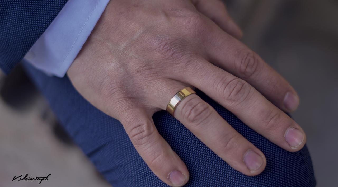 Męska dłoń z złotą obrączką na palcu serdecznym, widoczna faktura niebieskiego garnituru i mankiet koszuli.