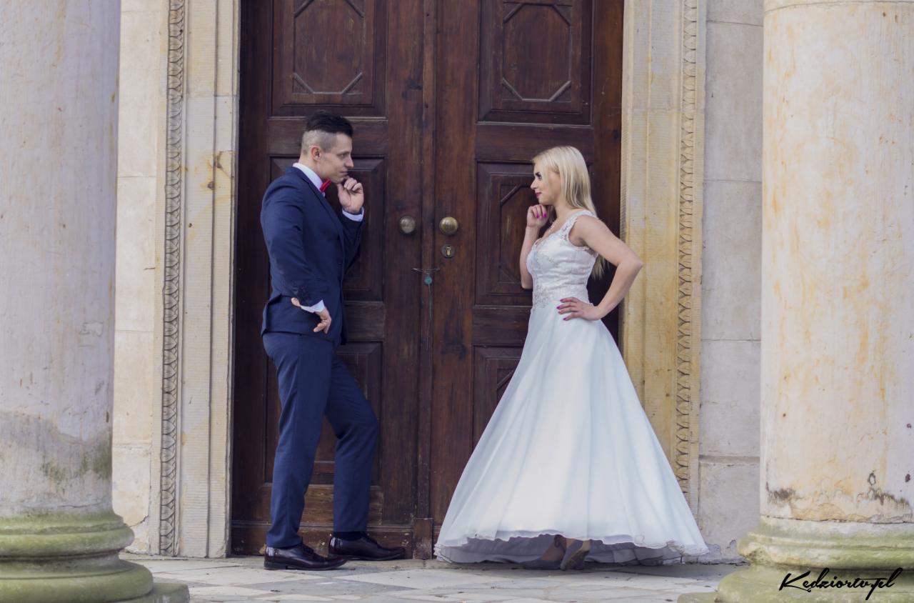 Para młoda: Panna Młoda w białej sukni i Pan Młody w granatowym garniturze, stoją naprzeciwko siebie, dotykając palcem brody, przed masywnymi, drewnianymi drzwiami z kolumnami.