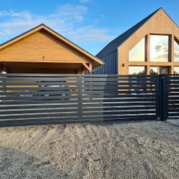 Nowoczesna, pozioma, przesuwna brama wjazdowa w kolorze antracytowym, z widocznym domem z dużymi oknami i drewnianą wiatą garażową w tle, na żwirowym podłożu.