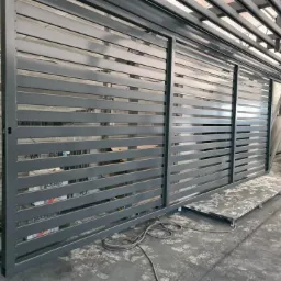 Szara, przesuwna brama przemysłowa z poziomymi szczeblami, widoczna w hali produkcyjnej z elementami wyposażenia.