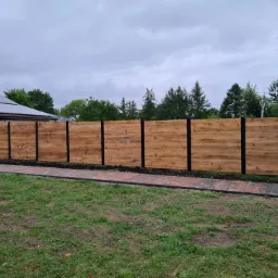 Drewniane ogrodzenie z czarnymi słupkami, widok z zewnątrz posesji, na tle domu z panelami słonecznymi i zieleni