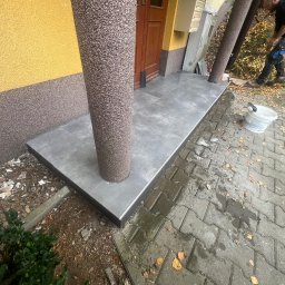 KOSBUD - Wykończone płytkami wejście do budynku z kolumnami. Widoczny fragment elewacji i pracownik w tle. Szare płytki z czarnym obramowaniem.