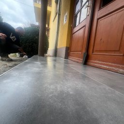 KOSBUD - Układanie szarych płytek na schodach wejściowych do domu. Fachowiec w trakcie pracy, widoczne narzędzia i fragment elewacji budynku z drewnianymi drzwiami.