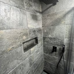 KOSBUD - Nowoczesna kabina prysznicowa z szarymi płytkami imitującymi kamień, wnęka na kosmetyki, czarna deszczownica i bateria. Minimalistyczny design.
