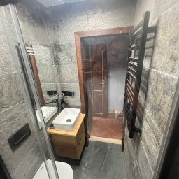 KOSBUD - Wykończona łazienka w Pielgrzymce z kabiną prysznicową, umywalką na drewnianej szafce i grzejnikiem drabinkowym na ścianie z szarych płytek imitujących kamień.