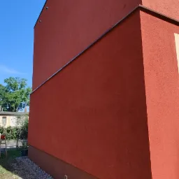 Czerwona ściana budynku z wykończonym tynkiem zewnętrznym, widoczne detale takie jak listwa okapowa, oświetlenie elewacyjne i cokół w innym odcieniu. U podstawy ściany ułożony drenaż z kamieni.