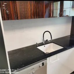 Nowoczesna kuchnia z czarnym, błyszczącym blatem, białą szafką i zlewem, oraz baterią z czarnym wężem. Nad blatem widoczna ściana wykończona gładzią oraz szafki z frontami w kolorze ciemnego drewna...