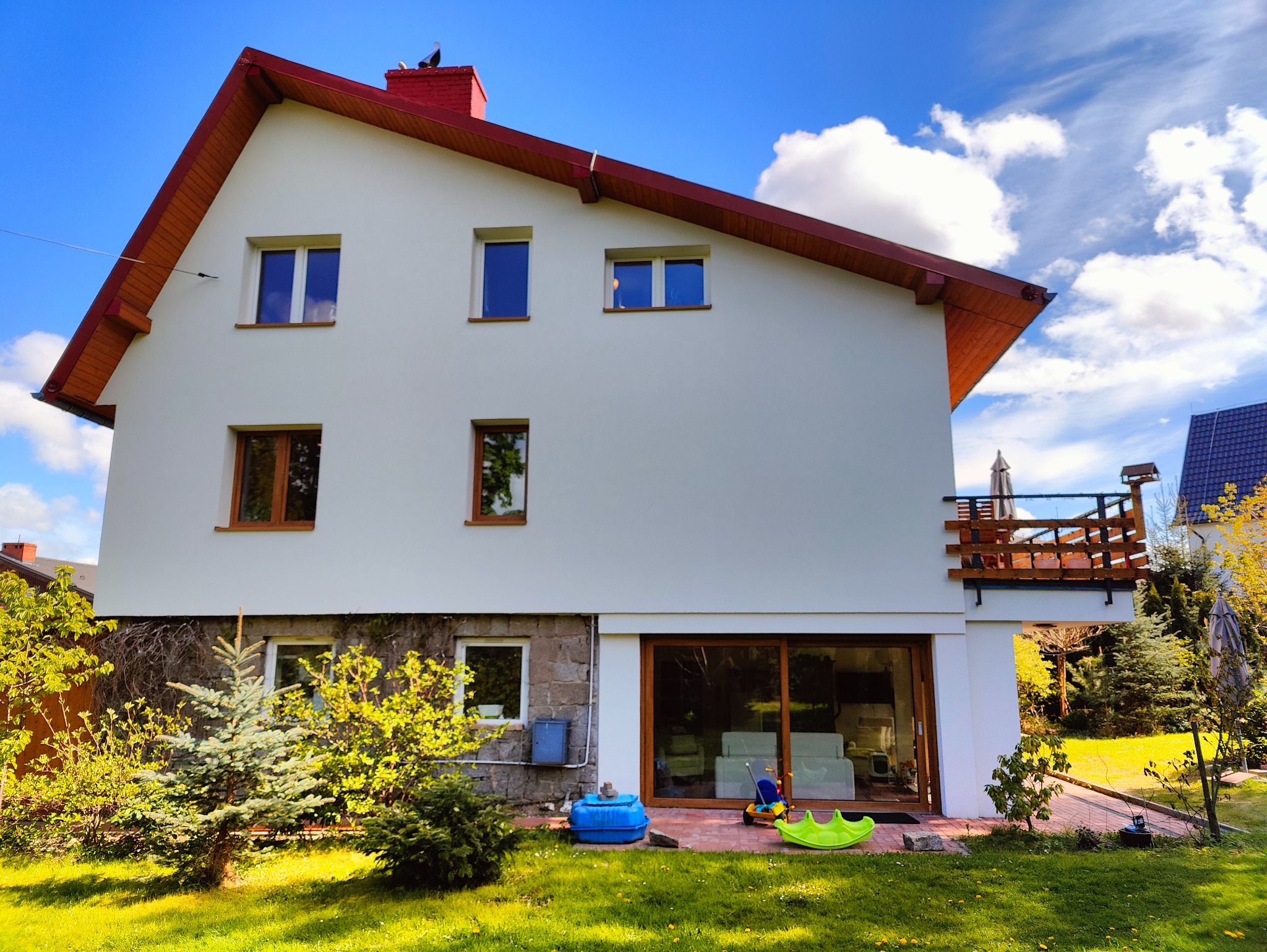 Dwukondygnacyjny dom z białymi ścianami, czerwonym dachem i drewnianymi oknami, otoczony zielonym trawnikiem i drzewami. Widoczny balkon z drewnianą balustradą.