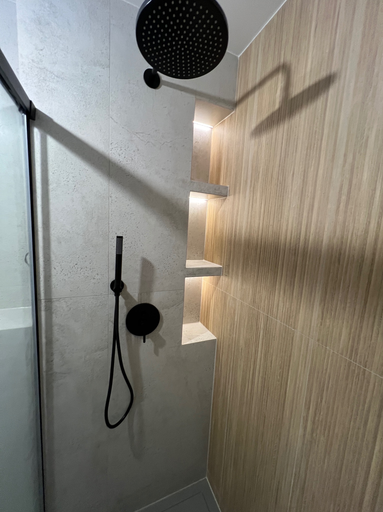 Nowoczesna kabina prysznicowa z czarną armaturą, jasnymi płytkami i wnęką z półkami podświetlonymi ciepłym światłem. Minimalistyczny design, eleganckie wykończenie.