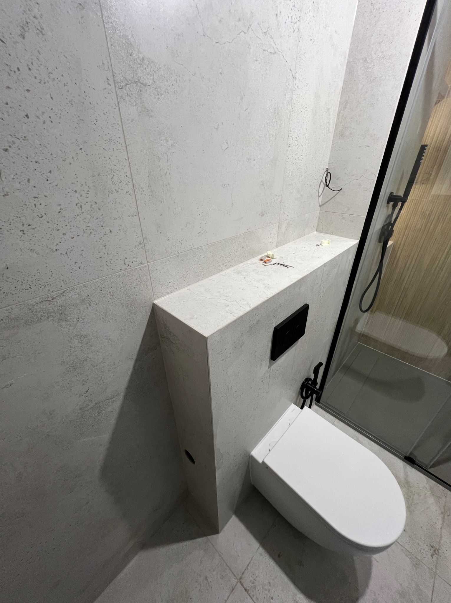 Wykończona łazienka z podwieszaną toaletą, zabudową z płytek imitujących kamień i czarną armaturą. Widoczna kabina prysznicowa z czarnymi profilami.