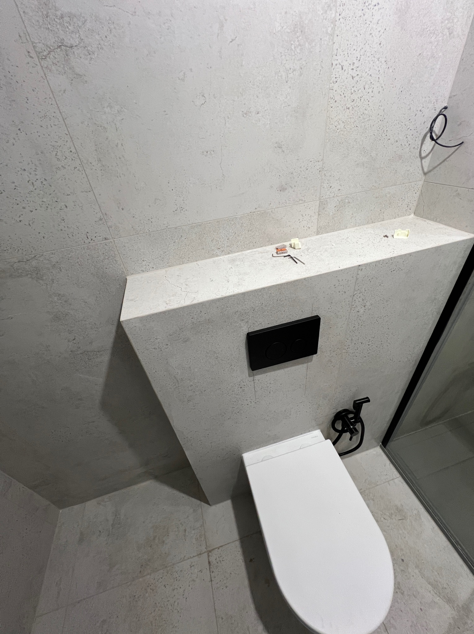 Nowoczesna toaleta podwieszana z czarną armaturą i przyciskiem spłukującym na tle szarych, teksturowanych płytek. Widoczne narzędzia i elementy instalacji na półce.