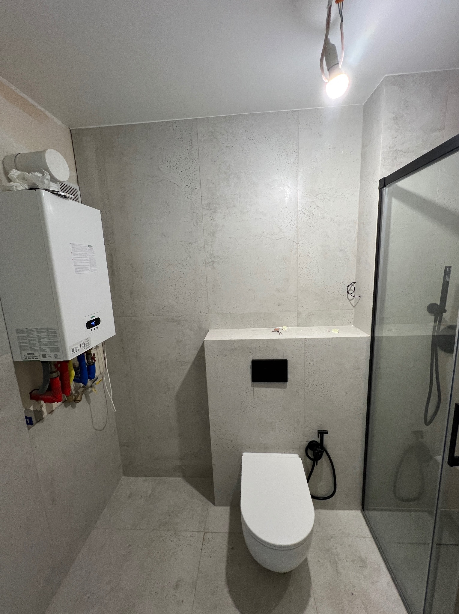 Nowoczesna łazienka z wiszącą toaletą, kabiną prysznicową i piecem gazowym. Płytki w odcieniach szarości. Minimalistyczny design. Widoczne instalacje.