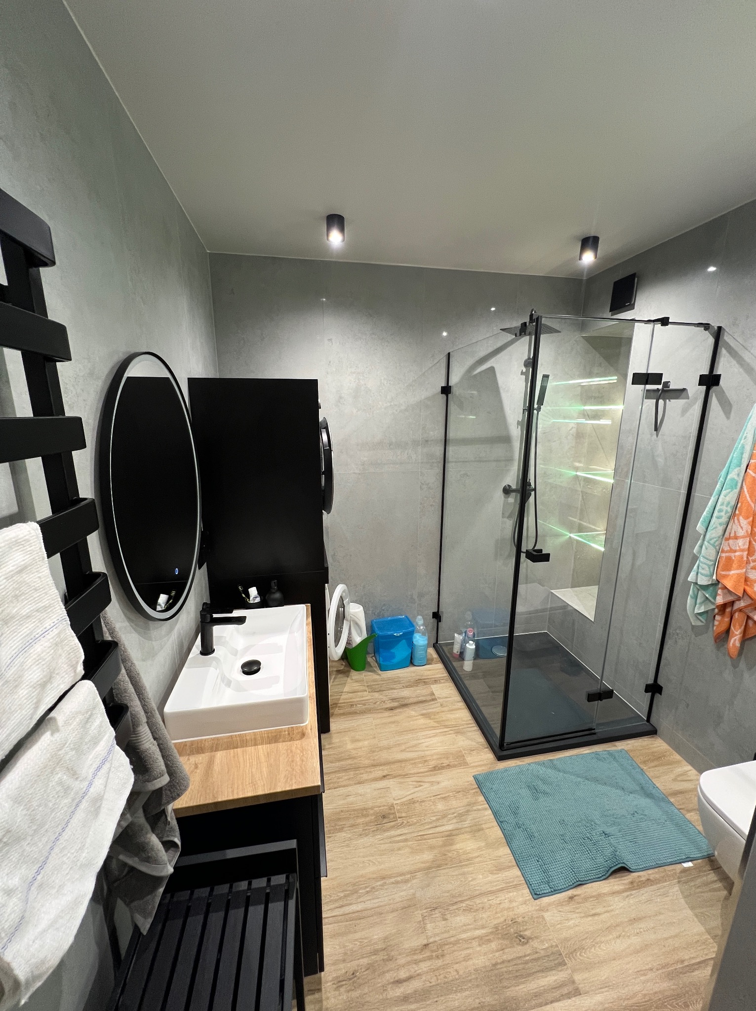 Nowoczesna łazienka z szarą glazurą, kabiną prysznicową typu walk-in z czarnymi ramami, umywalką nablatową i drewnopodobną podłogą. Minimalistyczny design.