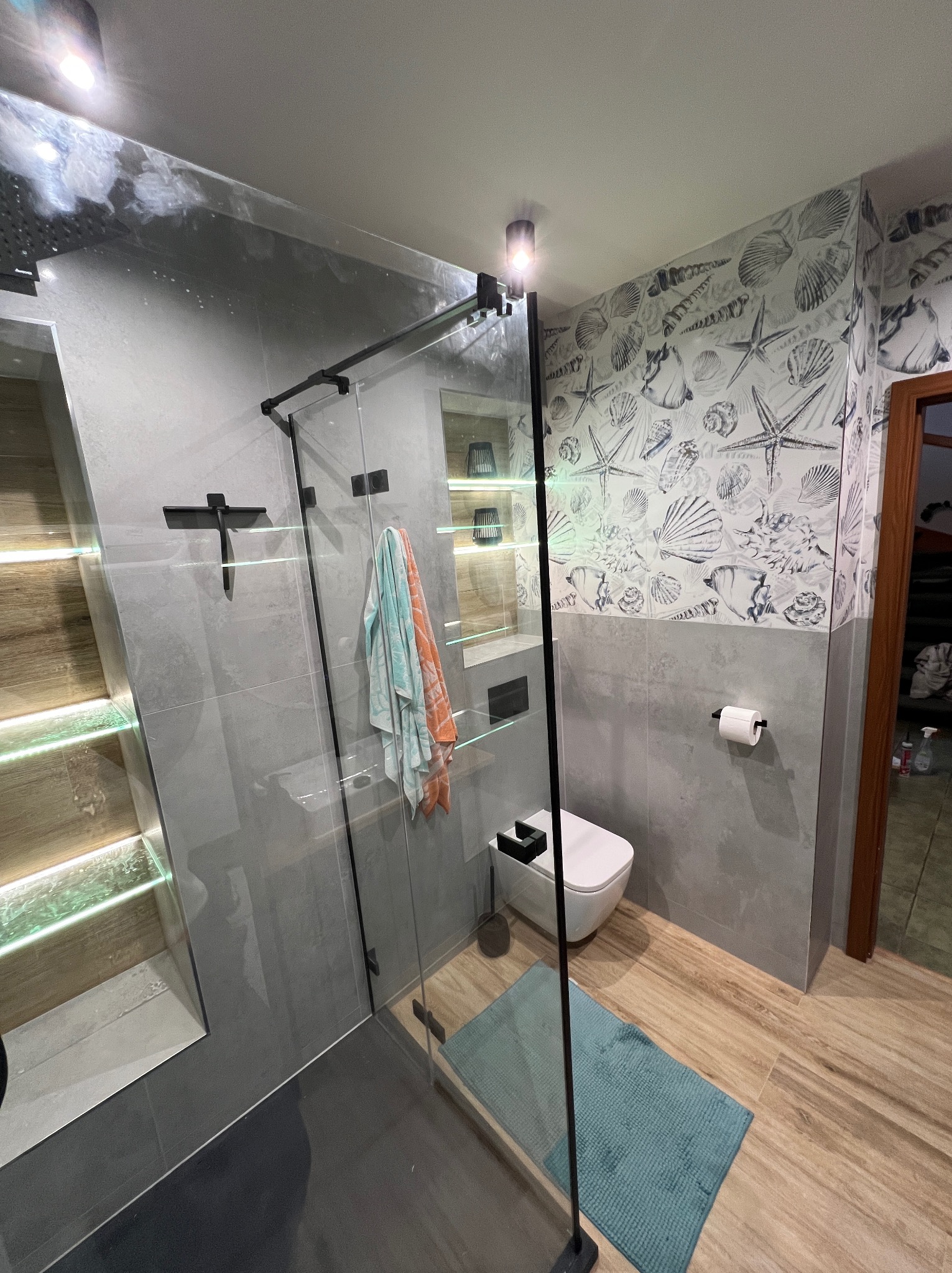 Nowoczesna łazienka z kabiną typu walk-in, szare płytki, akcenty drewna i tapeta w muszle. Oświetlenie LED podkreśla półki w strefie prysznica.