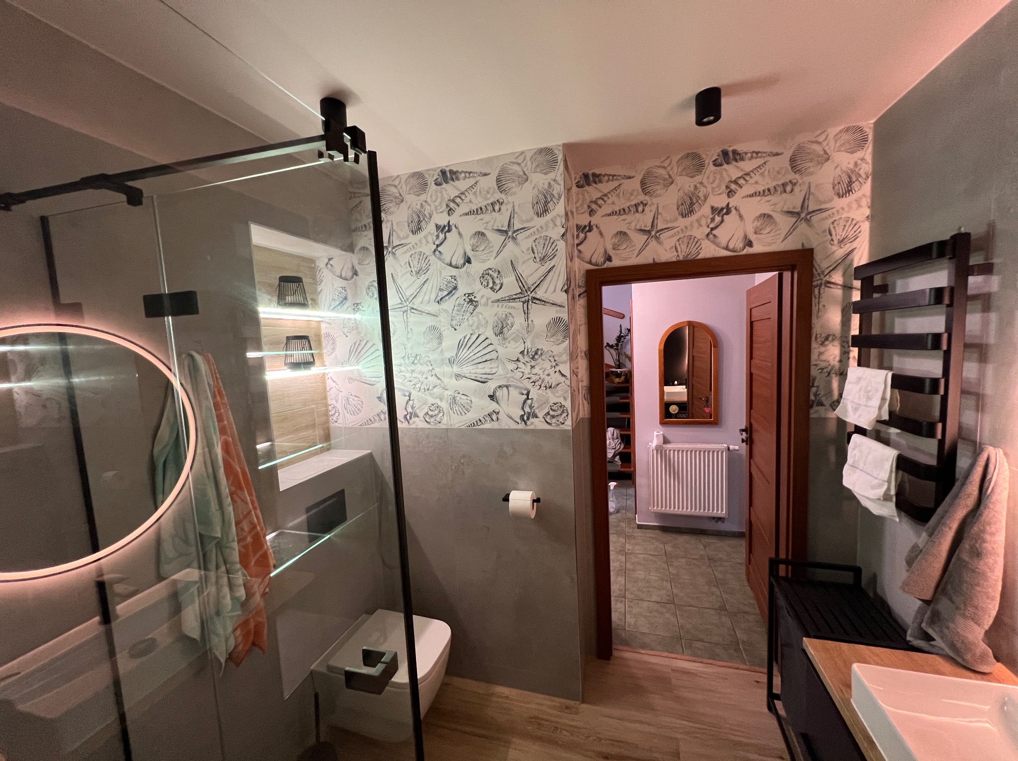 Nowoczesna łazienka z kabiną prysznicową typu walk-in, okrągłym lustrem z podświetleniem LED i tapetą w motywy morskie. Widoczny grzejnik drabinkowy i drewnopodobna podłoga.