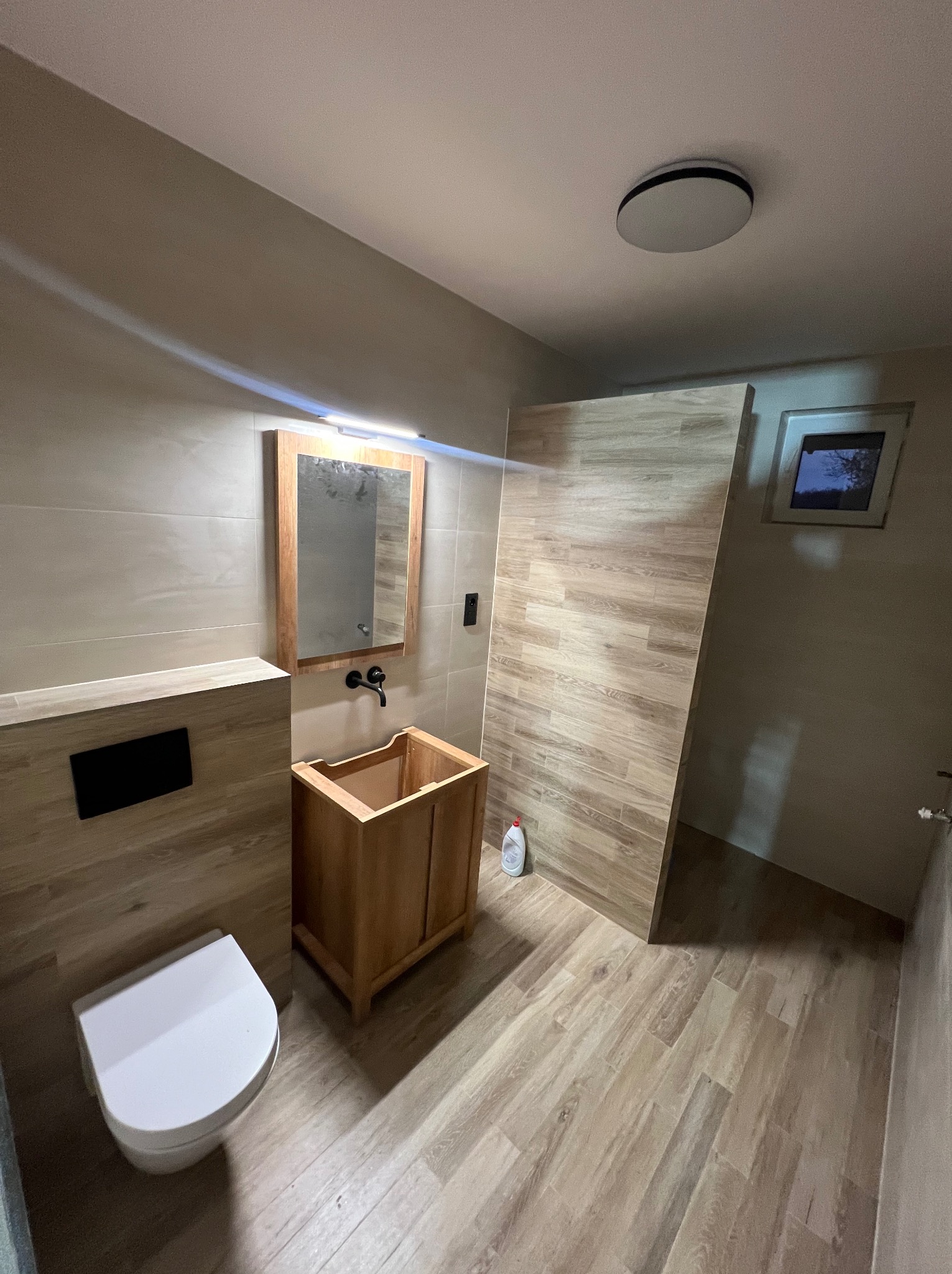 Nowoczesna łazienka w trakcie wykańczania: drewniana szafka pod umywalkę bez blatu, toaleta, lustro z oświetleniem i płytki imitujące drewno na ścianach i podłodze.