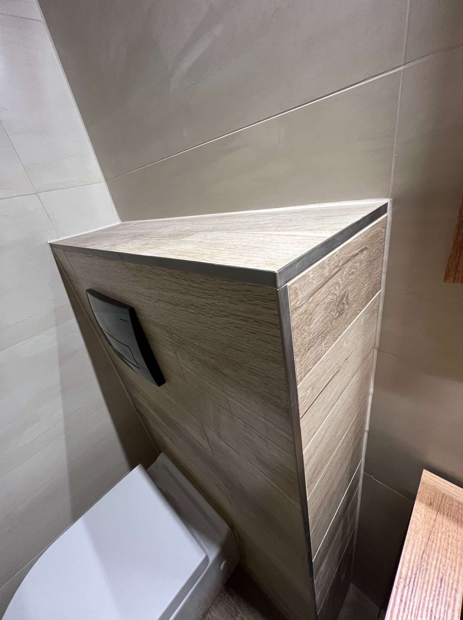 Kompakt WC zabudowany płytkami imitującymi drewno, z widocznym przyciskiem spłukującym i fragmentem miski. Całość w jasnej, nowoczesnej łazience z beżowymi płytkami.